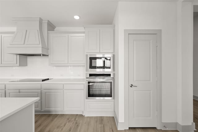 kitchen with light wood-type flooring, backsplash, stainless steel oven, custom exhaust hood, and white cabinets