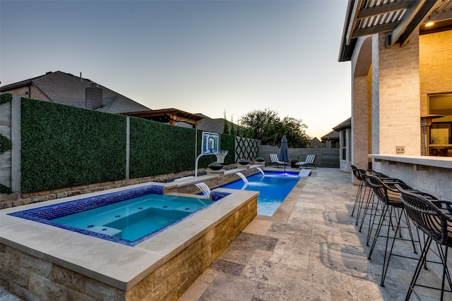 pool at dusk featuring an in ground hot tub, pool water feature, exterior bar, and a patio