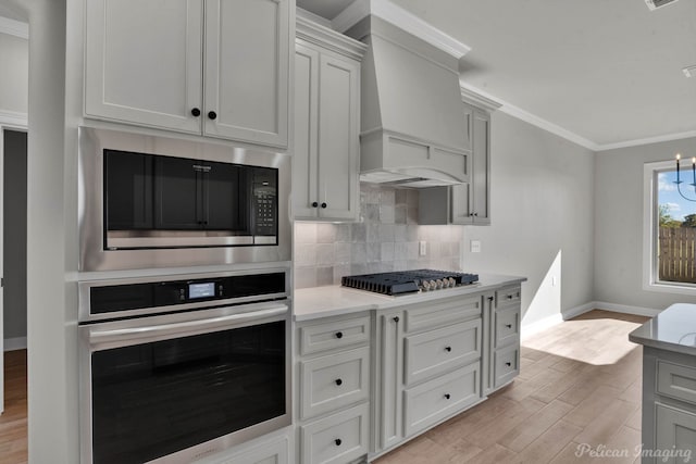 kitchen featuring appliances with stainless steel finishes, ornamental molding, custom range hood, a chandelier, and light hardwood / wood-style floors