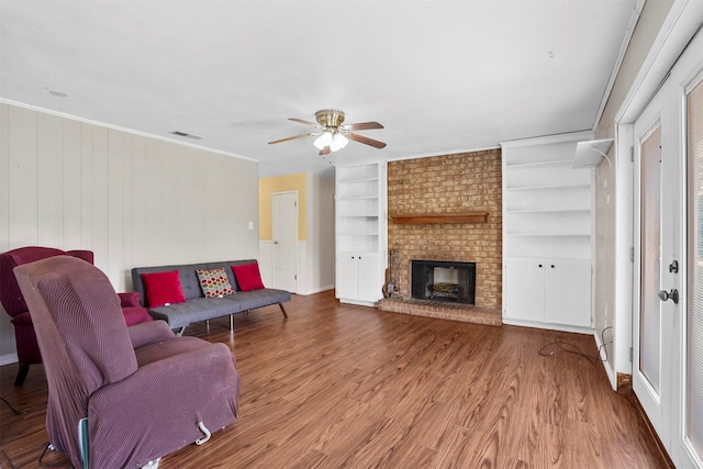 living room with built in shelves, ceiling fan, a brick fireplace, crown molding, and wood-type flooring