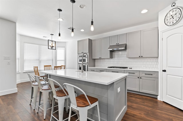 kitchen with hanging light fixtures, stainless steel appliances, gray cabinetry, a center island with sink, and dark hardwood / wood-style flooring