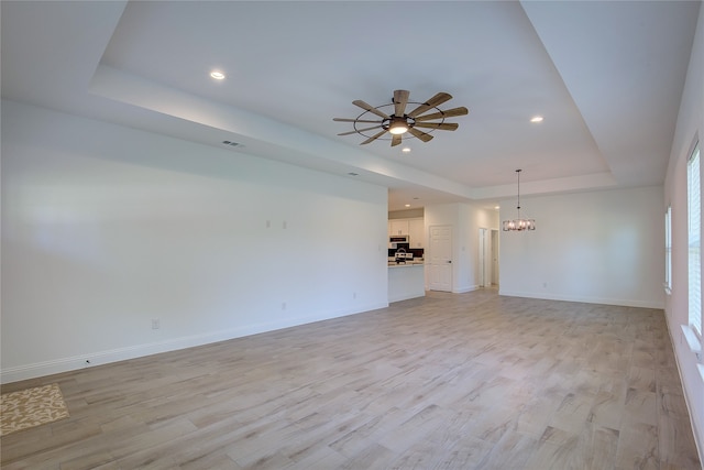 unfurnished living room with ceiling fan with notable chandelier, a tray ceiling, and light wood-type flooring