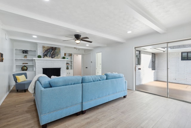 living room with ceiling fan, built in features, beam ceiling, a brick fireplace, and light hardwood / wood-style floors