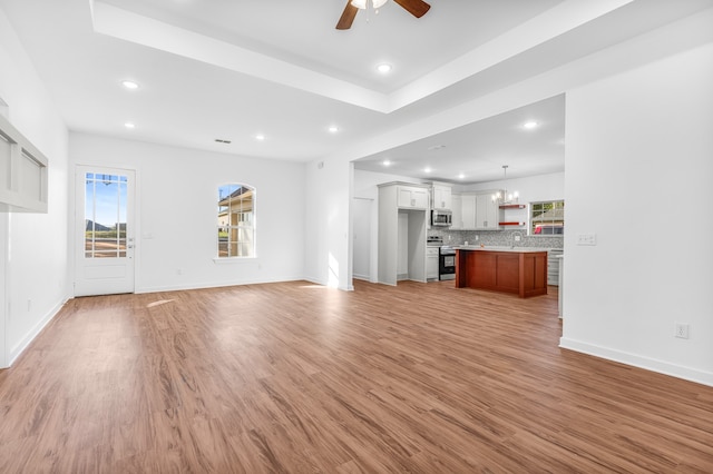 unfurnished living room featuring ceiling fan with notable chandelier, light hardwood / wood-style floors, and a tray ceiling