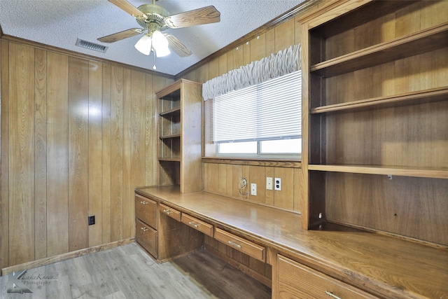 unfurnished office with wood walls, built in desk, ceiling fan, a textured ceiling, and light hardwood / wood-style flooring