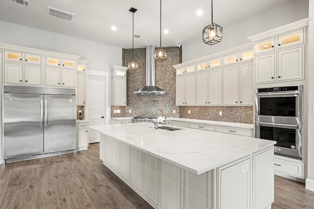 kitchen featuring wall chimney range hood, tasteful backsplash, an island with sink, pendant lighting, and stainless steel appliances