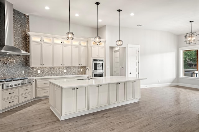 kitchen featuring a center island with sink, appliances with stainless steel finishes, light hardwood / wood-style floors, wall chimney exhaust hood, and sink