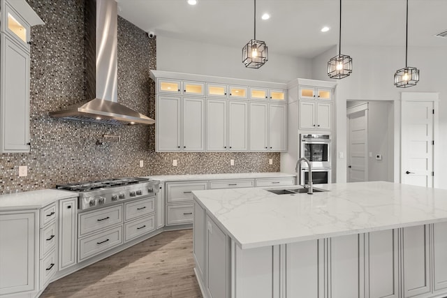 kitchen with wall chimney range hood, sink, a center island with sink, and white cabinets