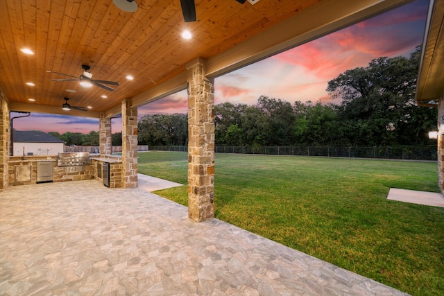 patio terrace at dusk with exterior kitchen, a yard, and ceiling fan