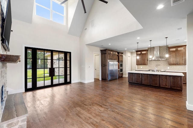 kitchen featuring wall chimney range hood, dark wood-type flooring, hanging light fixtures, stainless steel appliances, and a center island with sink