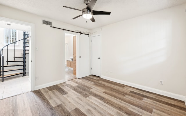 unfurnished bedroom with ensuite bathroom, a textured ceiling, ceiling fan, a barn door, and light hardwood / wood-style flooring