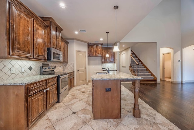 kitchen with light stone counters, pendant lighting, an island with sink, appliances with stainless steel finishes, and light hardwood / wood-style floors