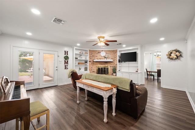 living room with a brick fireplace, built in features, ceiling fan, dark wood-type flooring, and ornamental molding