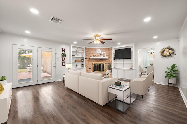 living room featuring dark wood-type flooring, ornamental molding, built in shelves, a brick fireplace, and ceiling fan with notable chandelier