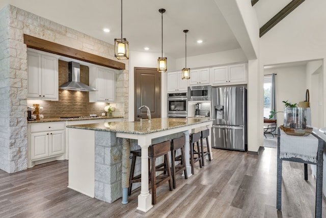 kitchen featuring stone countertops, a kitchen island with sink, wall chimney exhaust hood, white cabinetry, and appliances with stainless steel finishes