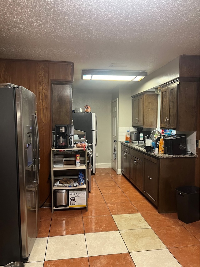 kitchen with stainless steel fridge, dark brown cabinets, light tile patterned flooring, and dark stone counters