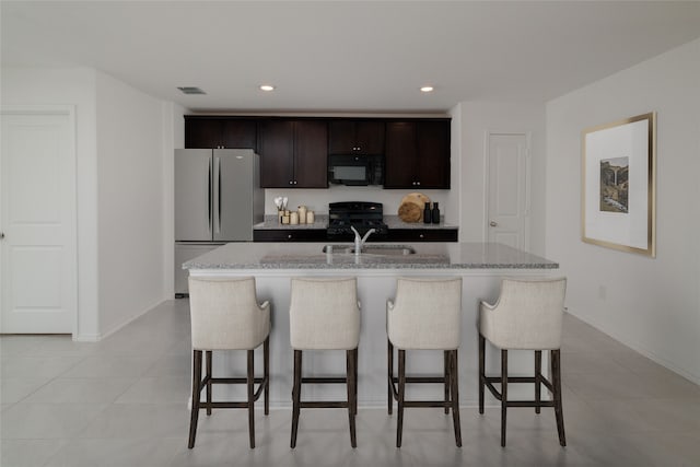 kitchen with dark brown cabinetry, light stone counters, a center island with sink, black appliances, and a breakfast bar