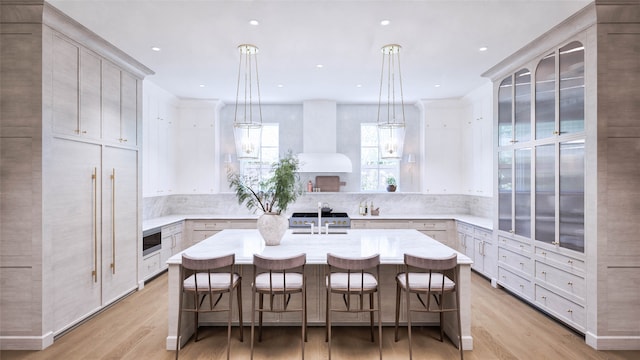 kitchen with decorative light fixtures, a center island with sink, wall chimney range hood, and light hardwood / wood-style flooring