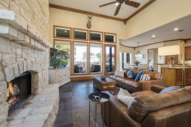 living room with a towering ceiling, sink, a stone fireplace, ceiling fan, and crown molding