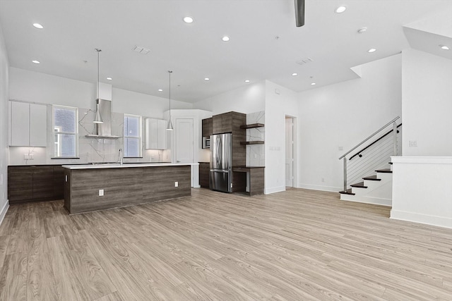 kitchen with light countertops, freestanding refrigerator, an island with sink, modern cabinets, and wall chimney exhaust hood