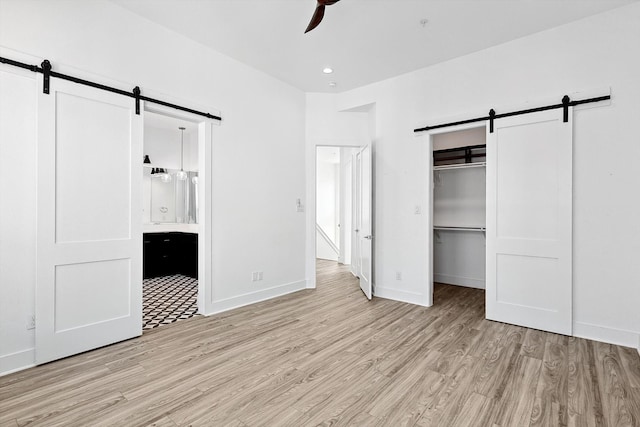 unfurnished bedroom featuring light wood-type flooring, ensuite bathroom, ceiling fan, a barn door, and a closet