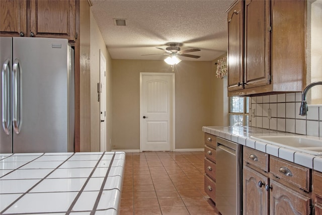 kitchen with a textured ceiling, stainless steel appliances, ceiling fan, sink, and tile counters