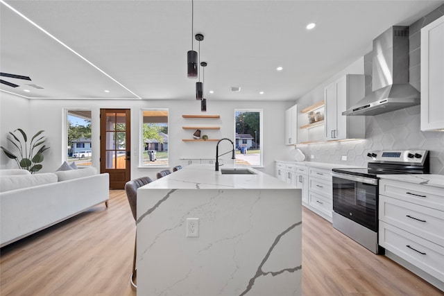 kitchen with white cabinets, hanging light fixtures, stainless steel electric range, a kitchen island with sink, and wall chimney range hood