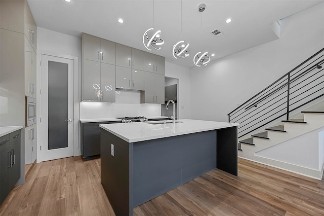 kitchen with an island with sink, light wood-type flooring, gray cabinetry, sink, and hanging light fixtures