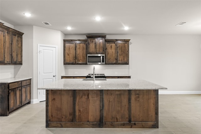 kitchen with dark brown cabinetry, sink, light stone counters, and a center island with sink