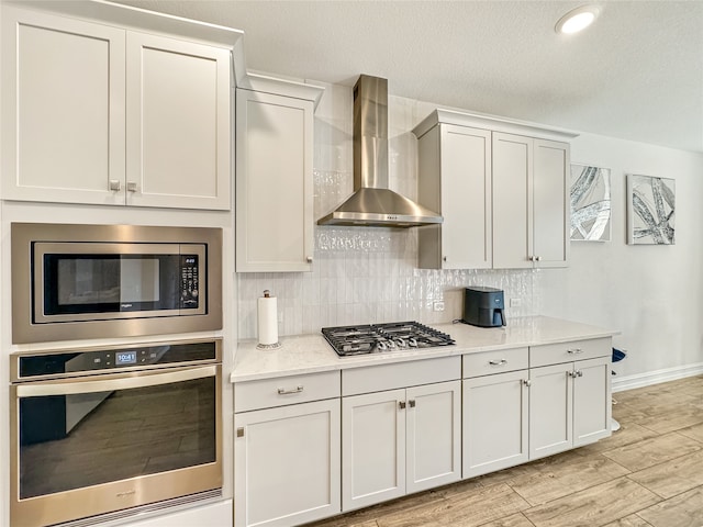 kitchen featuring light stone countertops, stainless steel appliances, wall chimney range hood, and light hardwood / wood-style flooring