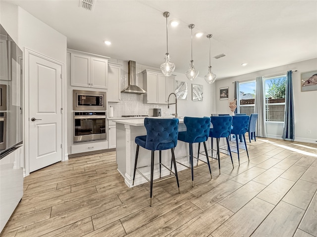 kitchen featuring decorative light fixtures, a center island with sink, wall chimney exhaust hood, white cabinetry, and stainless steel appliances