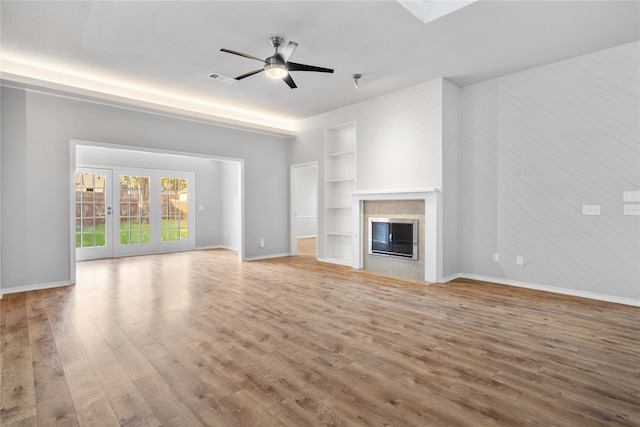 unfurnished living room featuring french doors, a tiled fireplace, built in features, light wood-type flooring, and ceiling fan