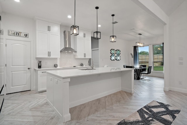 kitchen featuring wall chimney exhaust hood, a kitchen island with sink, tasteful backsplash, hanging light fixtures, and white cabinets