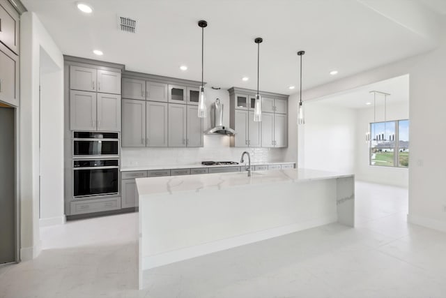 kitchen with light stone counters, a center island with sink, backsplash, and wall chimney range hood