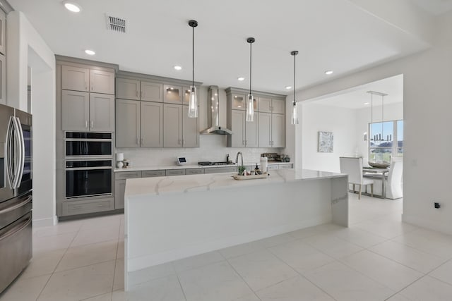 kitchen featuring a kitchen island with sink, light stone countertops, stainless steel appliances, wall chimney exhaust hood, and gray cabinetry
