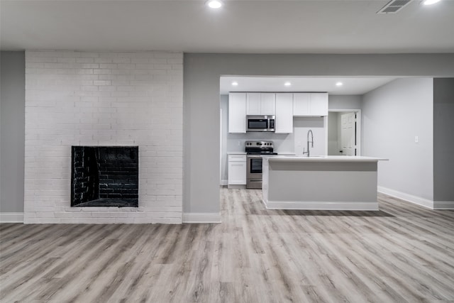 kitchen with a brick fireplace, light hardwood / wood-style floors, an island with sink, appliances with stainless steel finishes, and white cabinetry