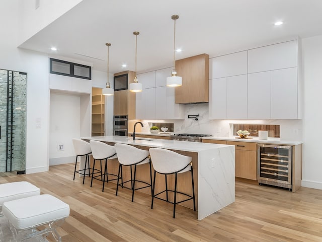 kitchen featuring beverage cooler, sink, a center island with sink, white cabinetry, and hanging light fixtures