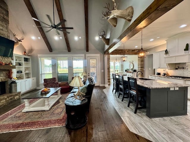 living room featuring ceiling fan with notable chandelier, light hardwood / wood-style flooring, sink, a stone fireplace, and vaulted ceiling with beams