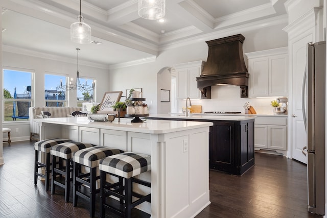 kitchen featuring white cabinetry, custom range hood, stainless steel refrigerator, hanging light fixtures, and a center island with sink