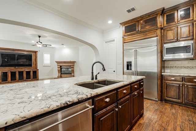 kitchen with sink, ornamental molding, built in appliances, light stone counters, and dark wood-type flooring