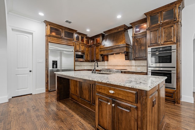 kitchen featuring sink, premium range hood, built in appliances, light stone countertops, and an island with sink