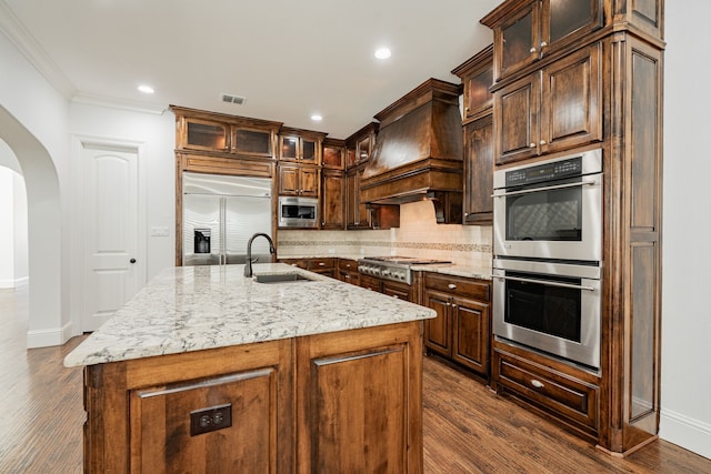 kitchen with sink, light stone counters, built in appliances, a center island with sink, and dark hardwood / wood-style flooring