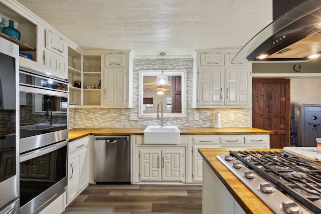 kitchen featuring butcher block counters, appliances with stainless steel finishes, wall chimney range hood, open shelves, and a sink