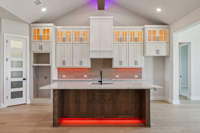 kitchen with an island with sink, light stone countertops, decorative backsplash, white cabinets, and vaulted ceiling