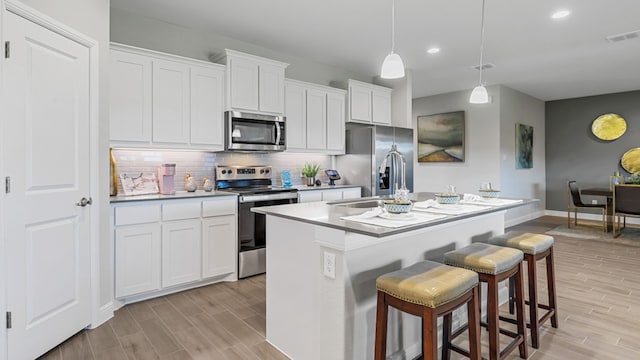 kitchen featuring hanging light fixtures, stainless steel appliances, a kitchen island with sink, white cabinets, and light wood-type flooring