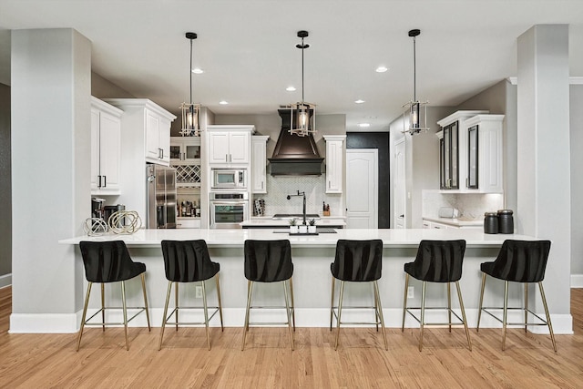 kitchen featuring pendant lighting, appliances with stainless steel finishes, white cabinetry, custom range hood, and a kitchen bar