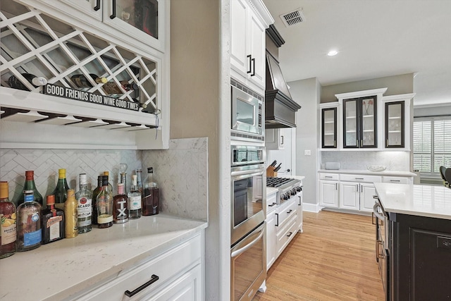 kitchen with premium range hood, appliances with stainless steel finishes, backsplash, white cabinets, and light wood-type flooring