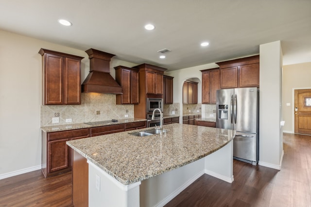 kitchen with a center island with sink, appliances with stainless steel finishes, custom range hood, light stone counters, and sink