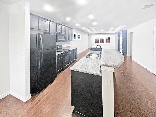 kitchen with light stone counters, a raised ceiling, sink, a center island with sink, and black appliances