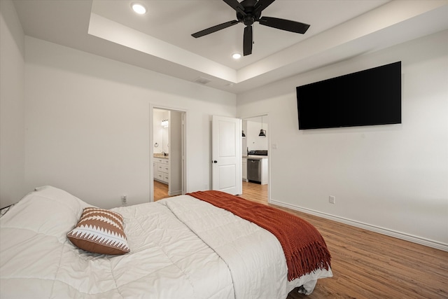 bedroom with light wood-type flooring, connected bathroom, a raised ceiling, and ceiling fan